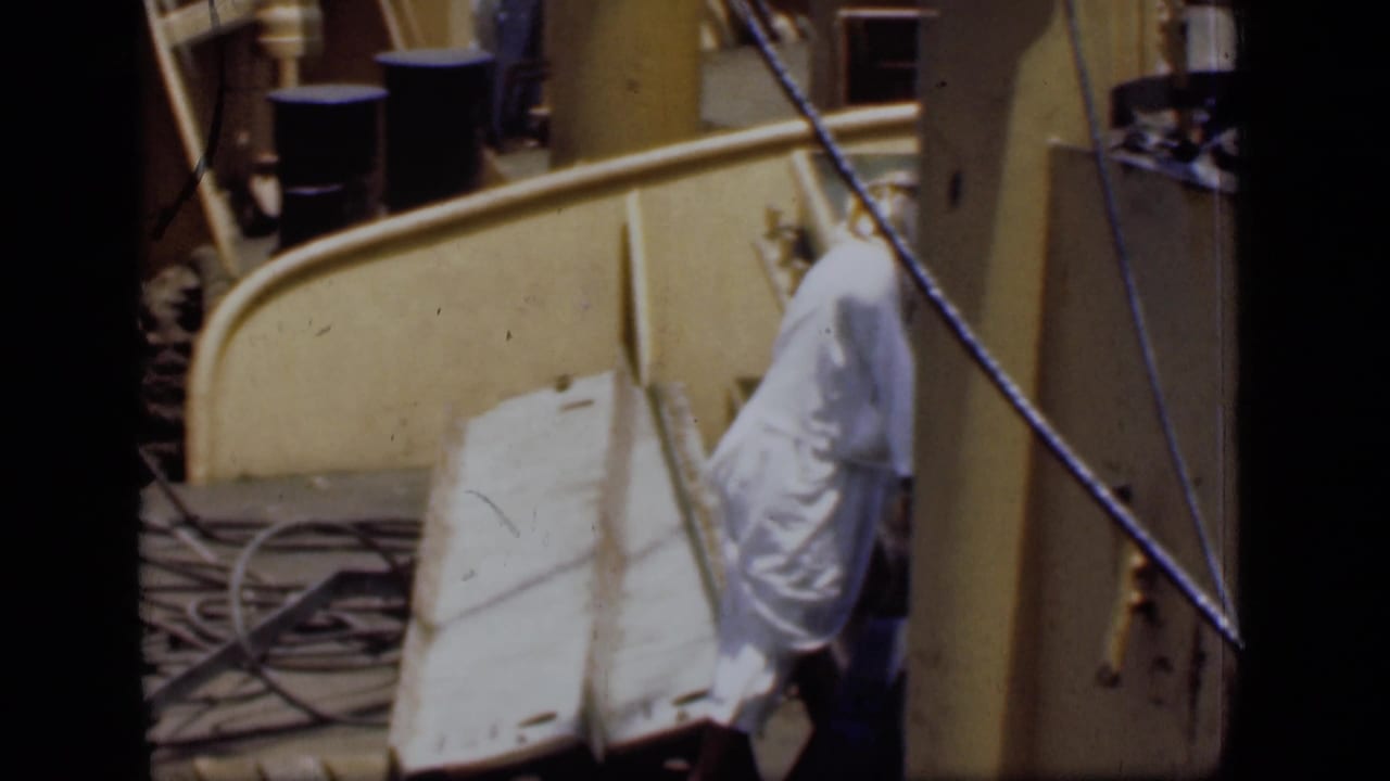 1946 Suez Canal frame with a man in white garments examining light-colored material beside ropes, barrels, and dock structures.