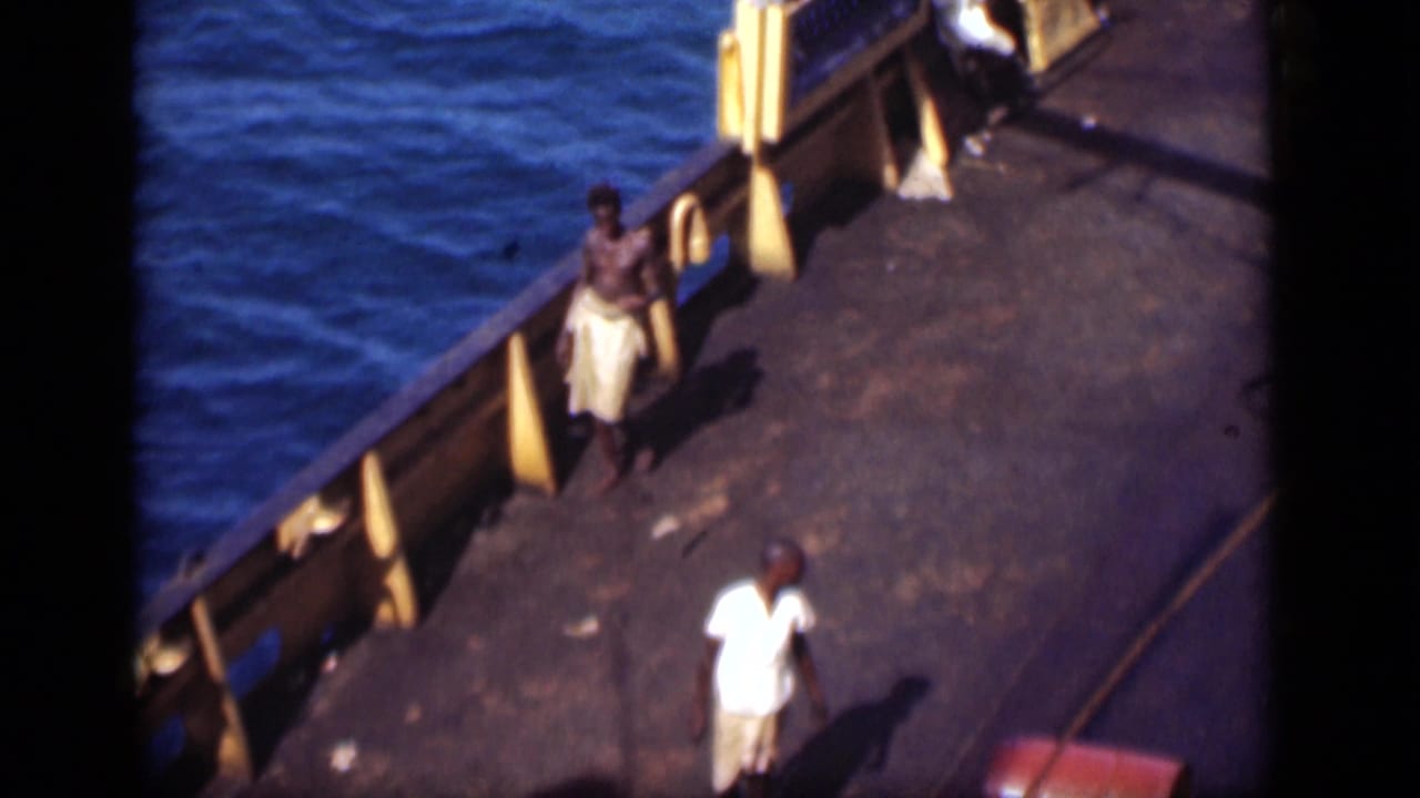 Two men walk on a boat deck beside blue canal water, one near the railing and one in a white shirt and shorts.