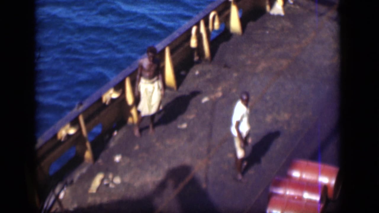 High-angle view of two men on a ship deck beside blue water and red barrels.