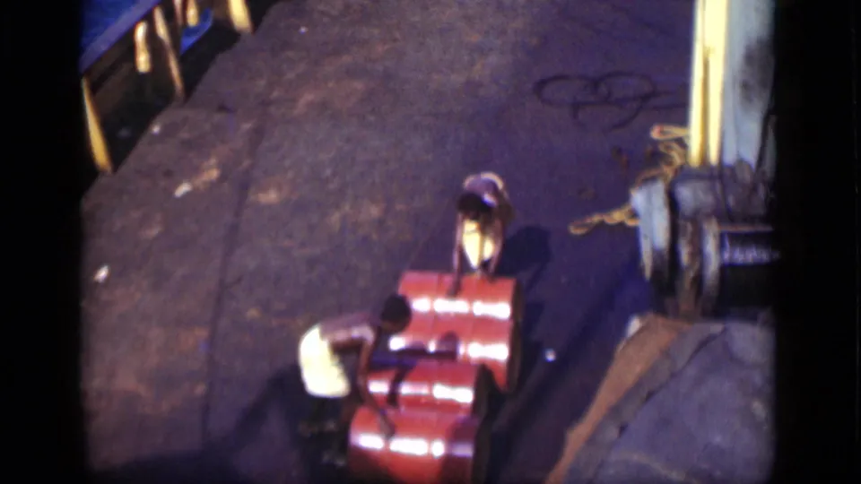 Two men on a 1946 Suez Canal vessel deck bend near bright red barrels, with rope and railings visible.