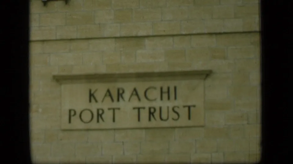 The 8mm frame shows a closer, slightly soft view of the Karachi Port Trust sign set into a stone wall.