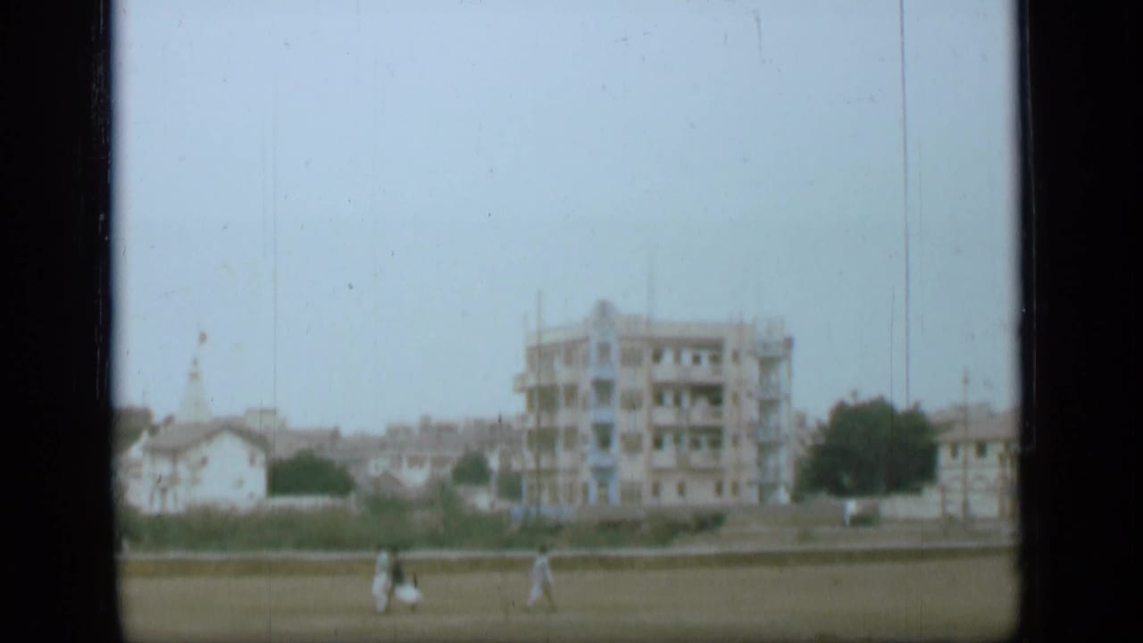 1946 8mm home-movie view associated with Suez Canal metadata, showing distant pedestrians crossing grass near low walls and urban buildings.