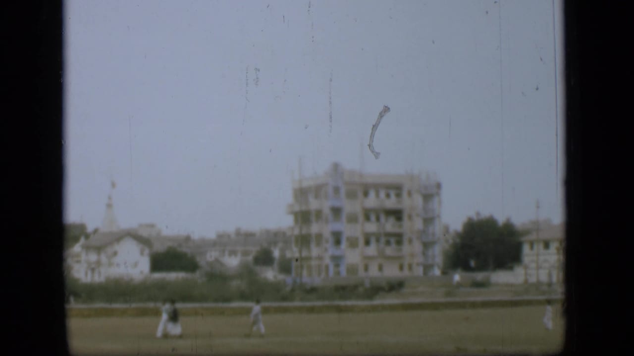 Distant people in light clothing walk across an open grassy area, with pale buildings, trees, and a larger structure beyond.
