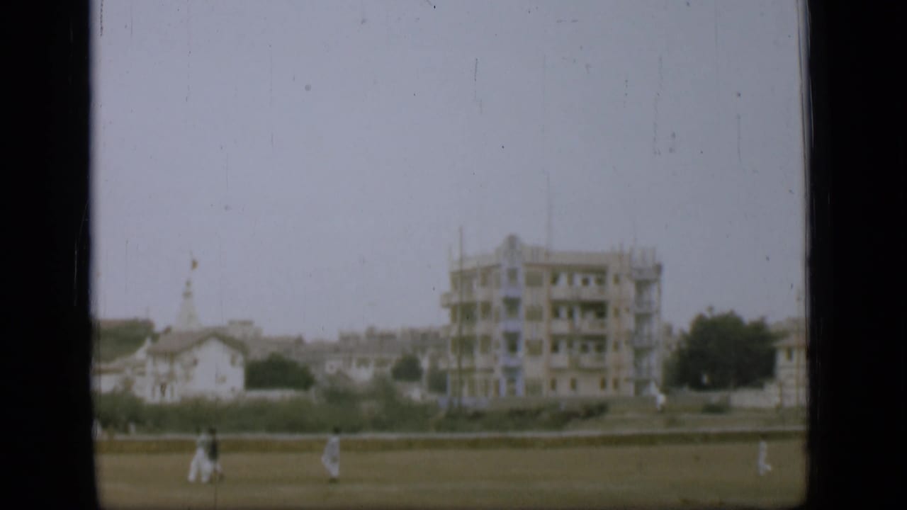 A soft 8mm frame shows distant pedestrians on grass, a low wall, pale houses, and a taller building with visible scaffolding.