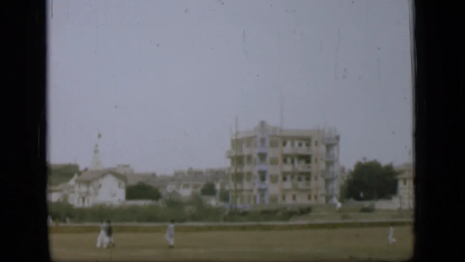 A soft 8mm frame shows distant pedestrians on grass, a low wall, pale houses, and a taller building with visible scaffolding.