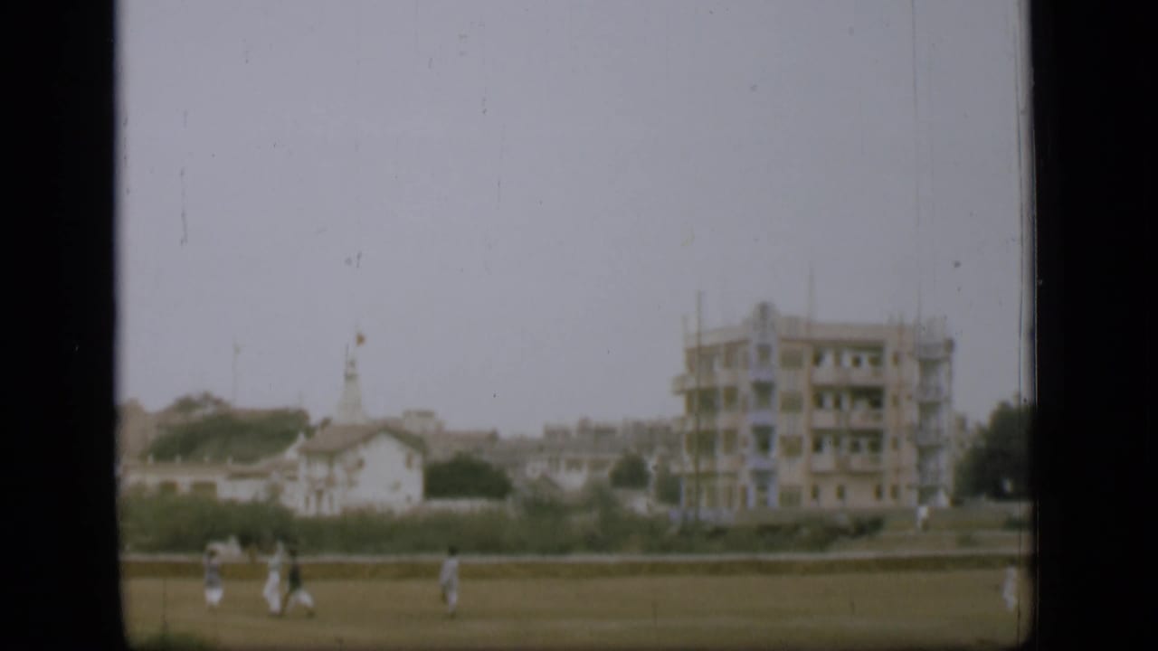 Distant figures move across grass in a 1946 8mm home-movie frame, with low buildings, trees, and a larger pale structure behind them.