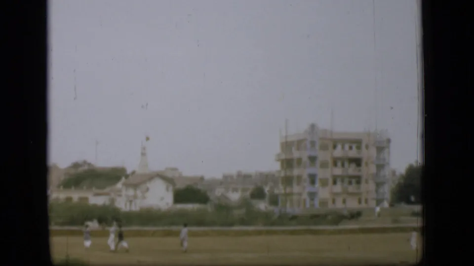 Distant figures move across grass in a 1946 8mm home-movie frame, with low buildings, trees, and a larger pale structure behind them.