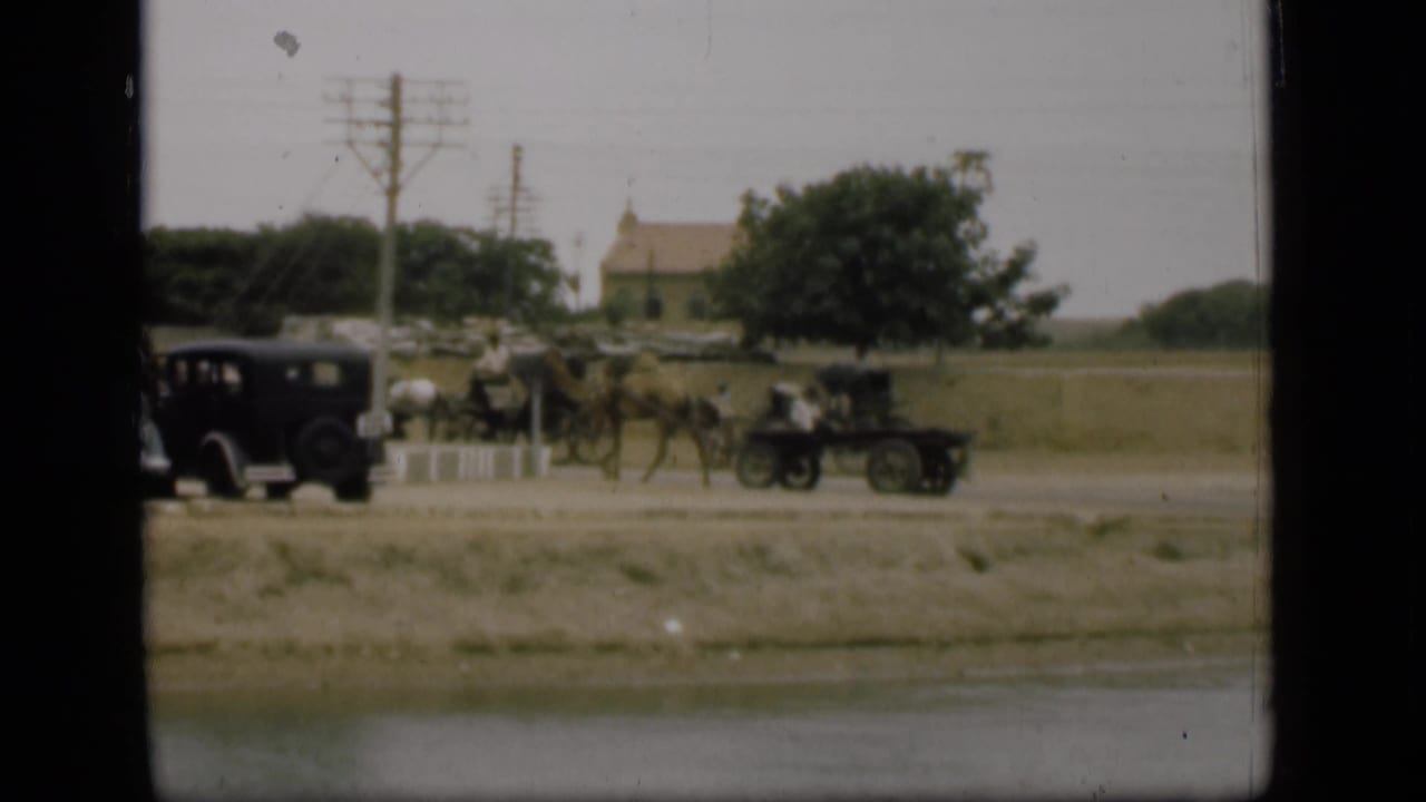 1946 Suez Canal area frame showing a camel-drawn cart, horse-drawn carriage, dark automobile, pedestrians, utility poles, and open land.