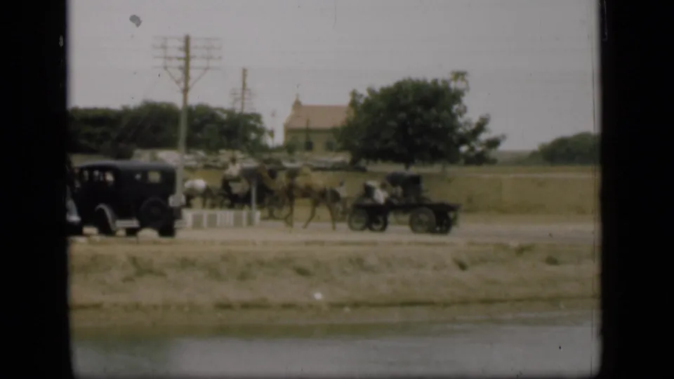 1946 Suez Canal area frame showing a camel-drawn cart, horse-drawn carriage, dark automobile, pedestrians, utility poles, and open land.