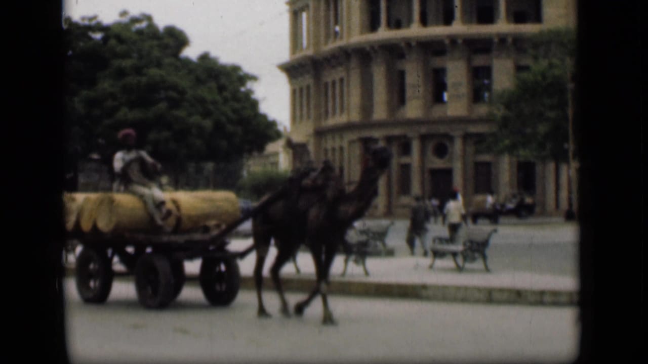 Camel-drawn cargo cart passes a large masonry building in the Suez Canal area, with a seated driver and pedestrians nearby.
