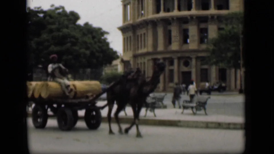 Camel-drawn cargo cart passes a large masonry building in the Suez Canal area, with a seated driver and pedestrians nearby.