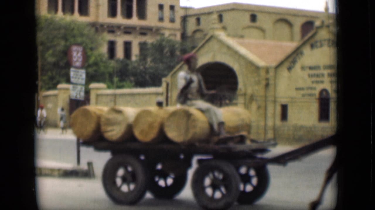 The camel-drawn cart passes a walled building entrance, with the driver seated above large cylindrical bundles in the stated Egyptian setting.