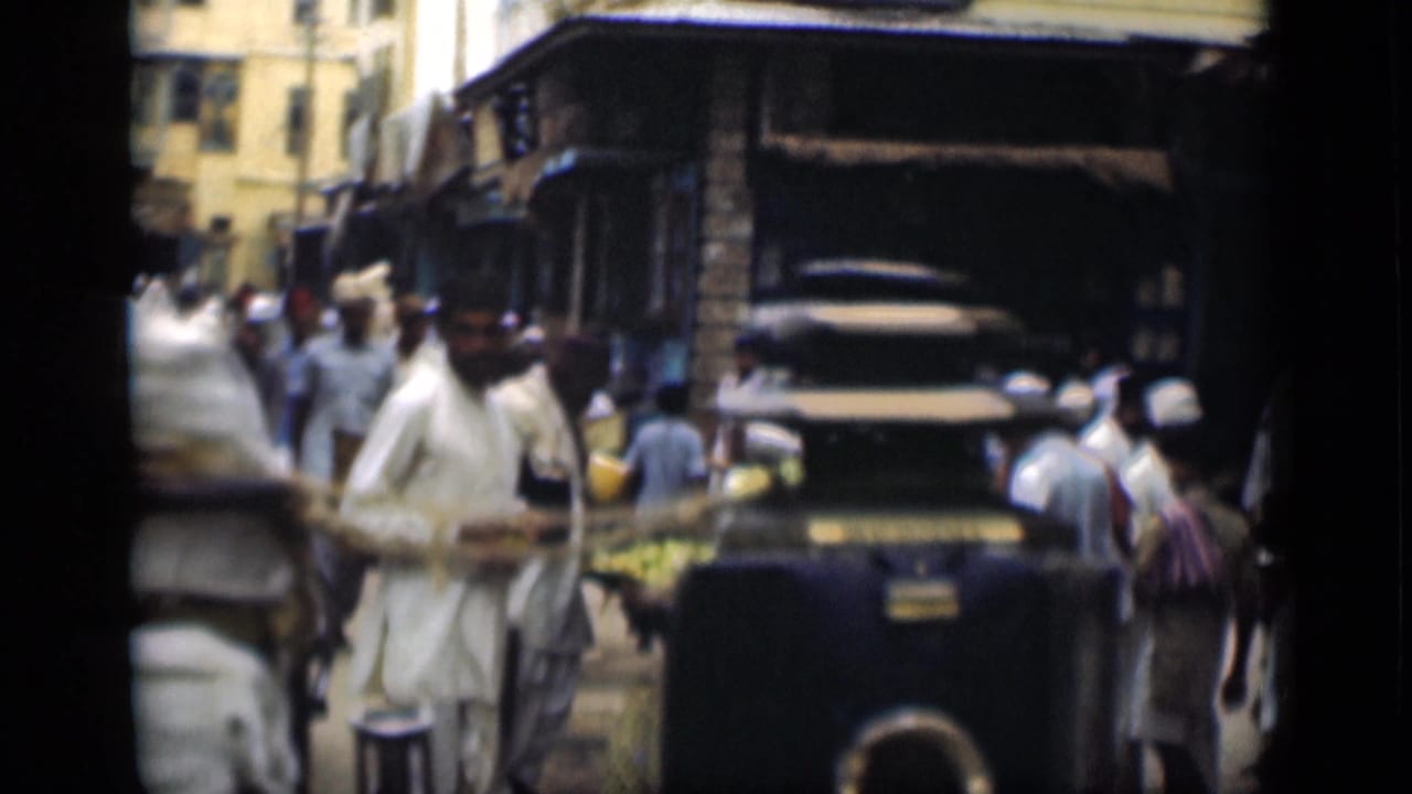 1946 Suez Canal, Egypt frame showing men in light robes and head coverings near a dark older car, market goods, awnings, and urban buildings.
