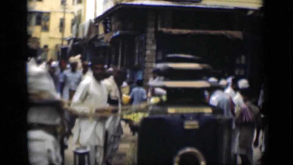1946 Suez Canal, Egypt frame showing men in light robes and head coverings near a dark older car, market goods, awnings, and urban buildings.