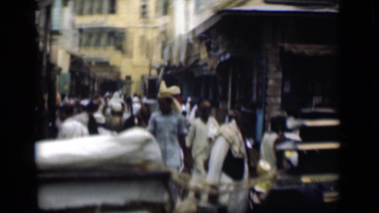 1946 Suez Canal, Egypt home-movie frame looking down a crowded street with pedestrians, a dark older car, shaded storefronts, and pale buildings.