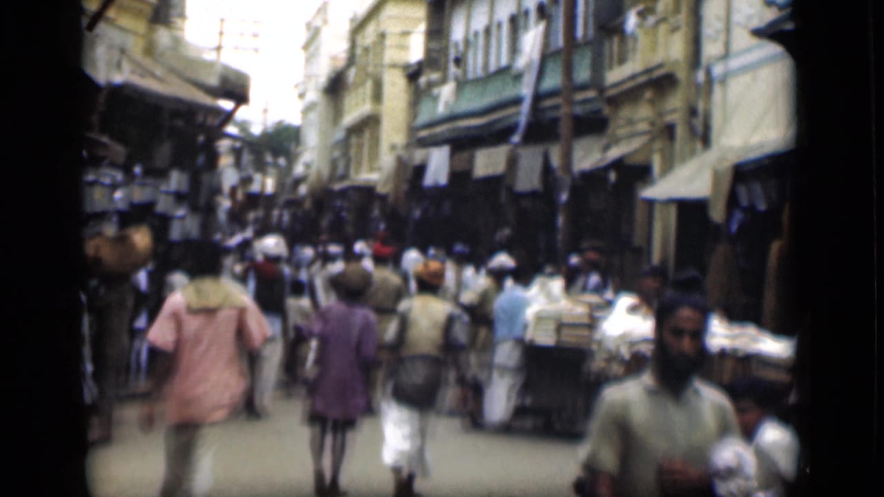 Handheld 8mm frame of a crowded Suez Canal region market street with carts, pedestrians, and shaded storefronts.