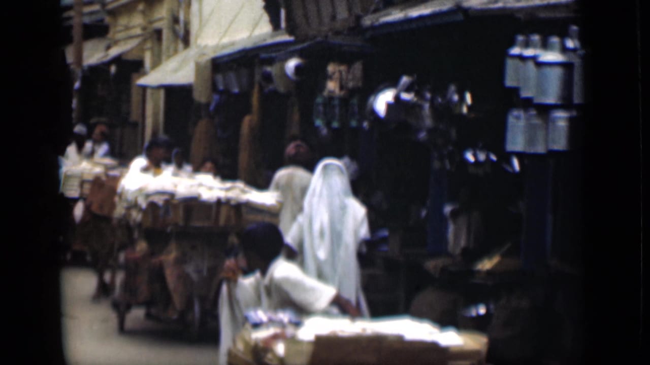Suez Canal region market lane in 1946 with pedestrians, a loaded cart, shopfronts, and a veiled figure seen from behind.
