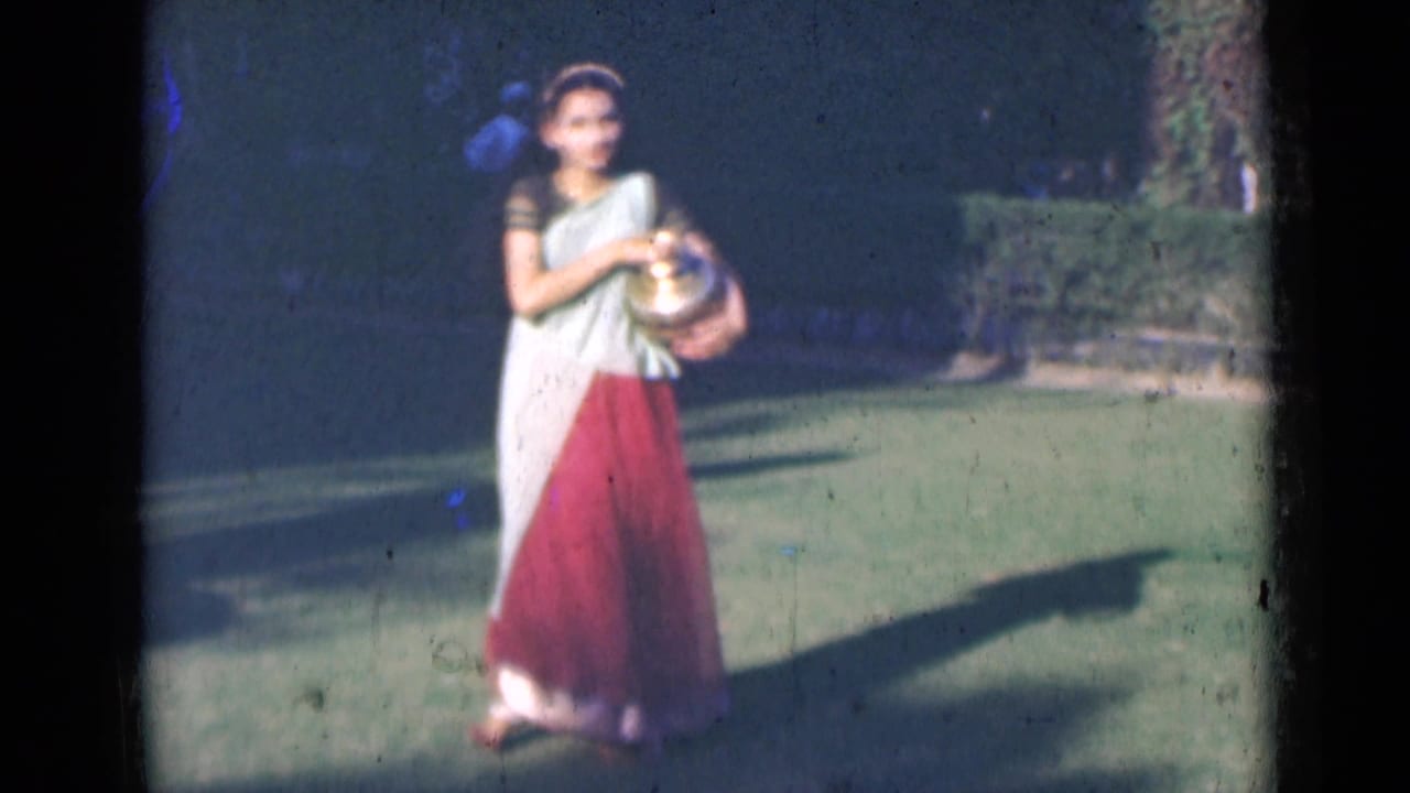In 1947 India home-movie footage, a young woman in a light and red saree holds a brass pot while walking on a garden lawn.