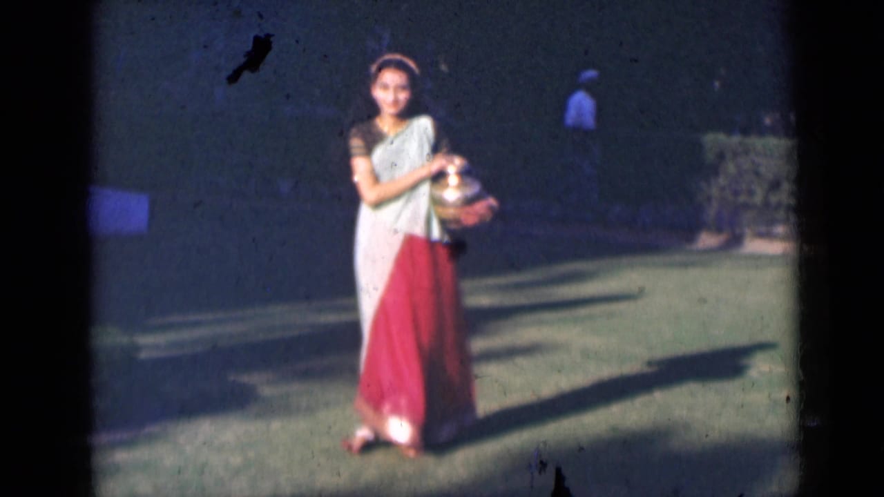 The woman faces the camera on a garden lawn in India, holding a polished brass pot as long shadows cross the grass.