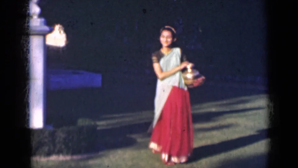 A young woman in a saree walks near a white pillar and lamp fixture, holding a brass pot in a shaded garden setting.