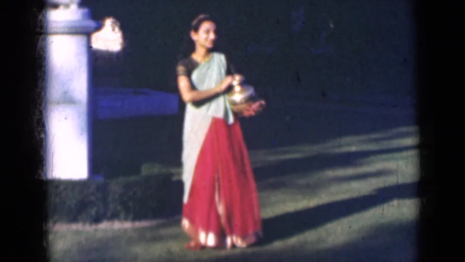The 8mm frame shows the woman in a saree standing with a brass pot beside a white pillar, with lawn and shadows behind her.