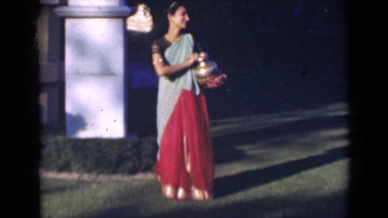 In 1947 India home-movie footage, a woman in a light green drape and red skirt holds a brass pot beside a white garden pillar.