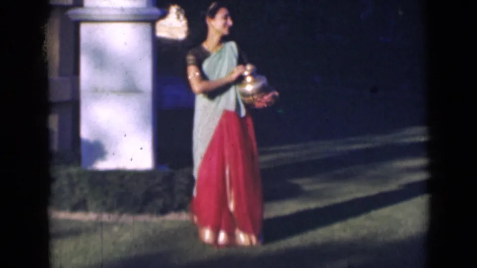 In 1947 India home-movie footage, a woman in a light green drape and red skirt holds a brass pot beside a white garden pillar.