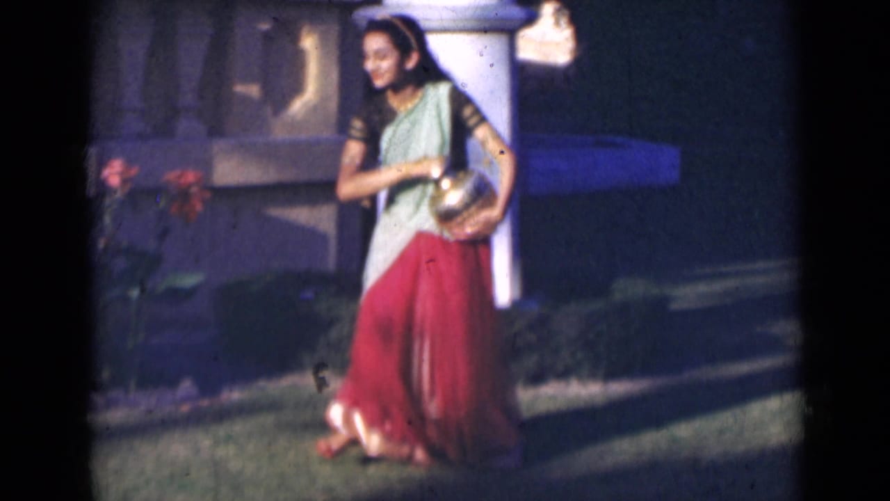 In 1947 India, the woman smiles while walking outdoors with a brass pot, framed by grass, flowers, and a pale pillar.