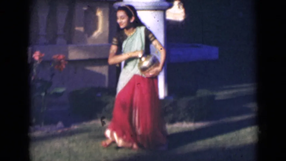 In 1947 India, the woman smiles while walking outdoors with a brass pot, framed by grass, flowers, and a pale pillar.