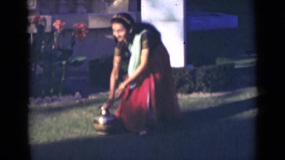 The woman bends down in the garden setting, appearing to set a brass pot on the grass during the brief 1947 India clip.