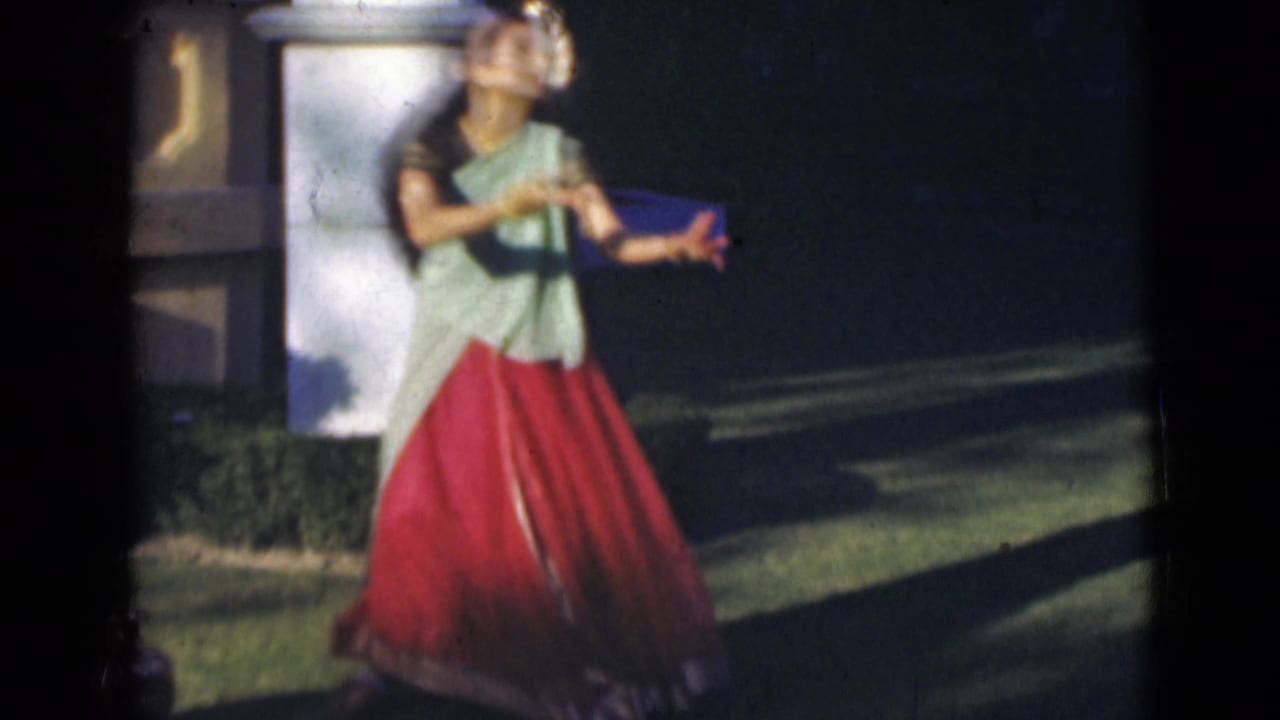 In 1947 India home-movie footage, a young woman in a green and red sari dances outdoors beside a white pillar and hedge.