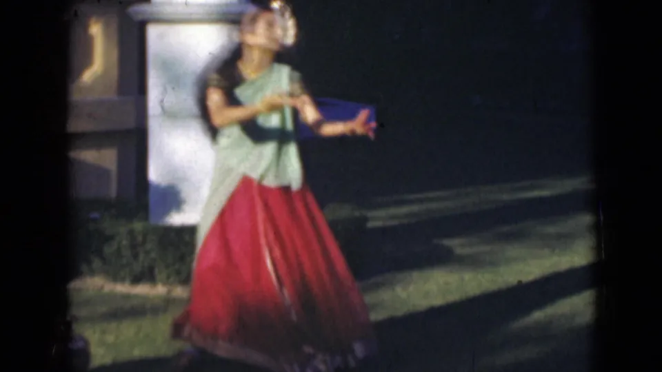 In 1947 India home-movie footage, a young woman in a green and red sari dances outdoors beside a white pillar and hedge.
