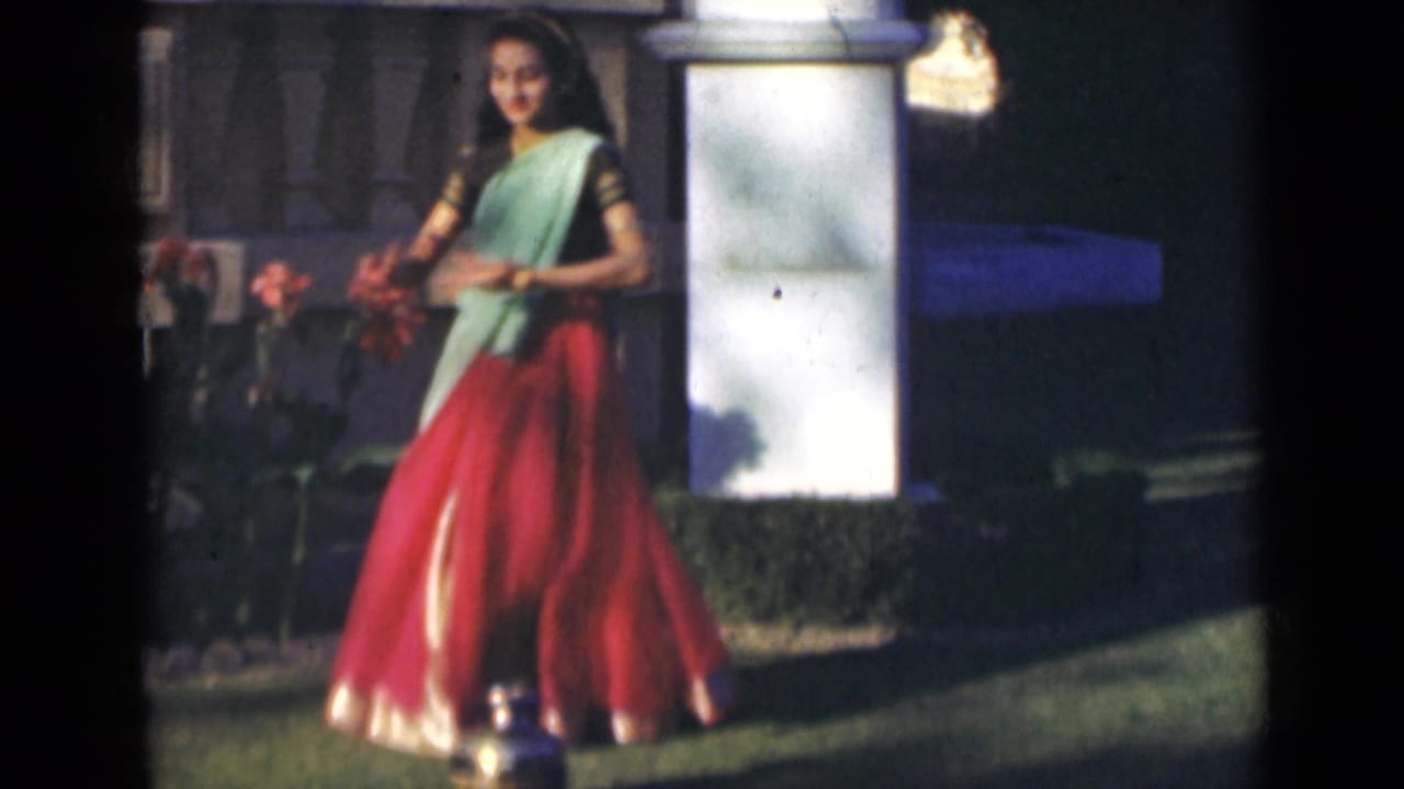 A young woman in a bright sari performs a dance pose in a garden setting in India, with flowers and a white pillar behind her.