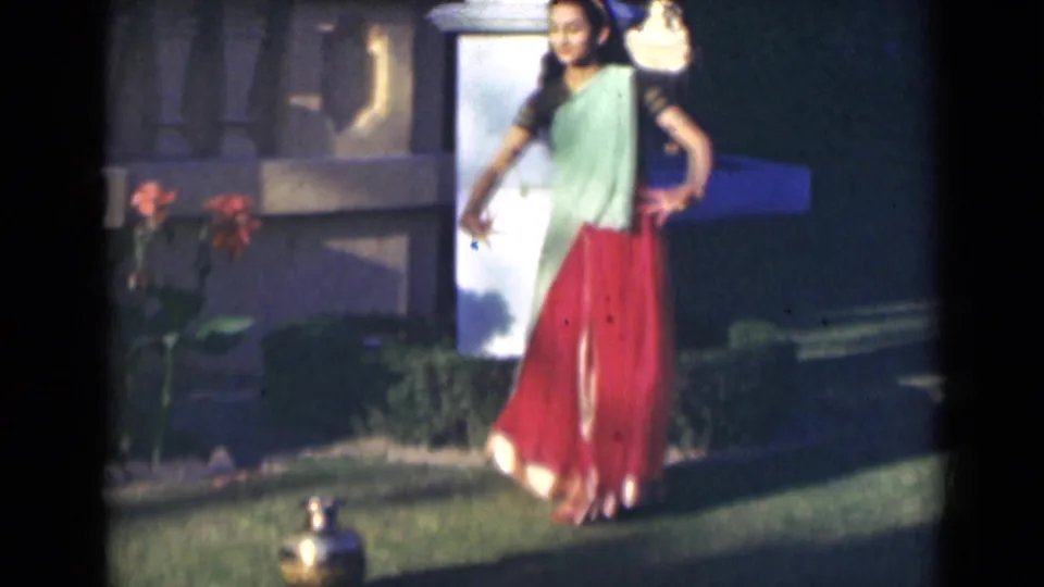 The 8mm frame shows the sari-clad dancer near a white pillar and flowers, with a small brass pot visible on the grass.