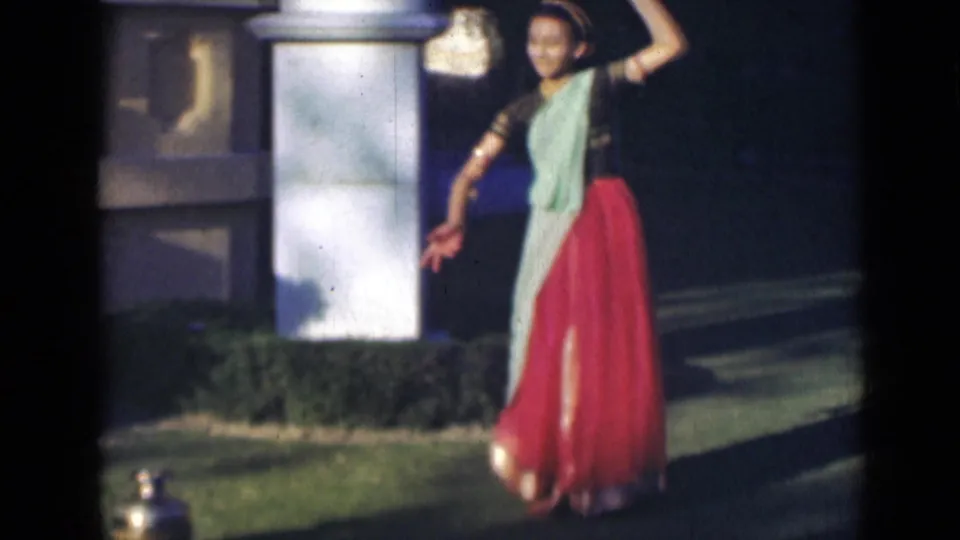 In the closing frame, the dancer raises one arm in a poised gesture beside a white pillar, hedge, and brass pot in the garden.