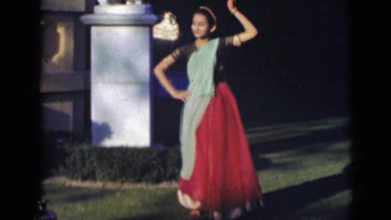 Young woman dancing outdoors in a red skirt and pale green drape beside a white pillar.