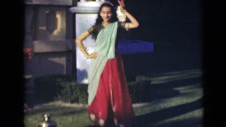 The dancer faces the camera in a garden setting, wearing a red skirt, pale green drape, and dark blouse in 1947 India home-movie footage.
