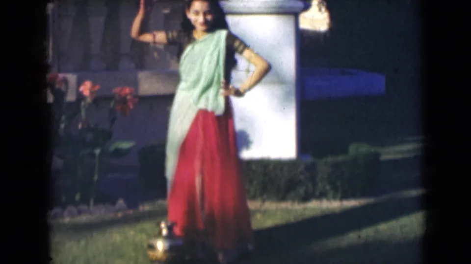 A young woman in traditional clothing moves through a dance pose outdoors, with flowers, grass, hedges, and a white pillar visible.