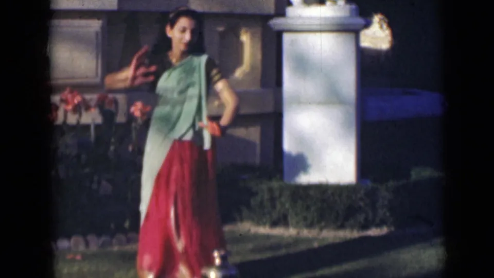 At the end of the clip, the young woman turns in a dance gesture near the pillar and hedges, with red fabric visible by her shoulder.