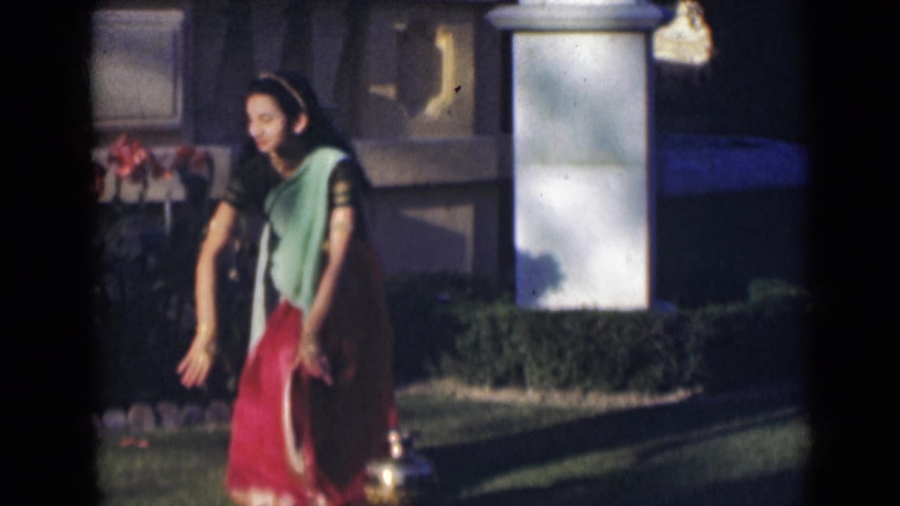 Woman in a green and red sari dancing on a garden lawn near flowers and a brass vessel.