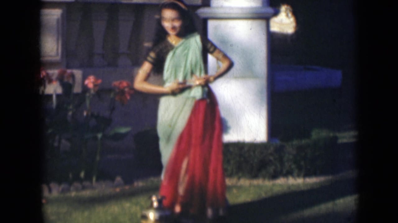 Woman in a sari standing mid-dance in a garden with flowers, columns, and a brass vessel nearby.