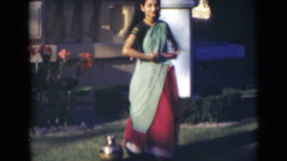The 1947 India home movie shows the woman moving through a dance step on the lawn beside flowers and a sunlit column.