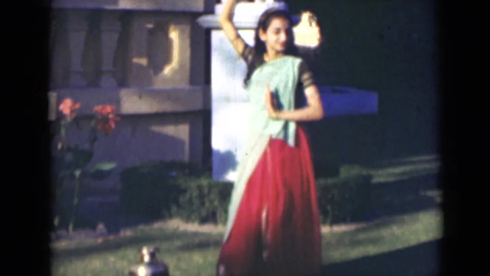 In a 1947 India garden scene, the woman raises one arm while dancing in a green and red sari near flowers and a brass vessel.