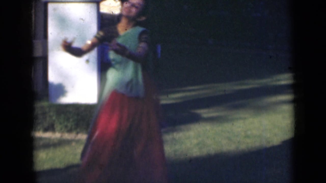 In 1947 India home-movie footage, a young woman in costume raises her arms while dancing outdoors beside a white pillar and lawn.