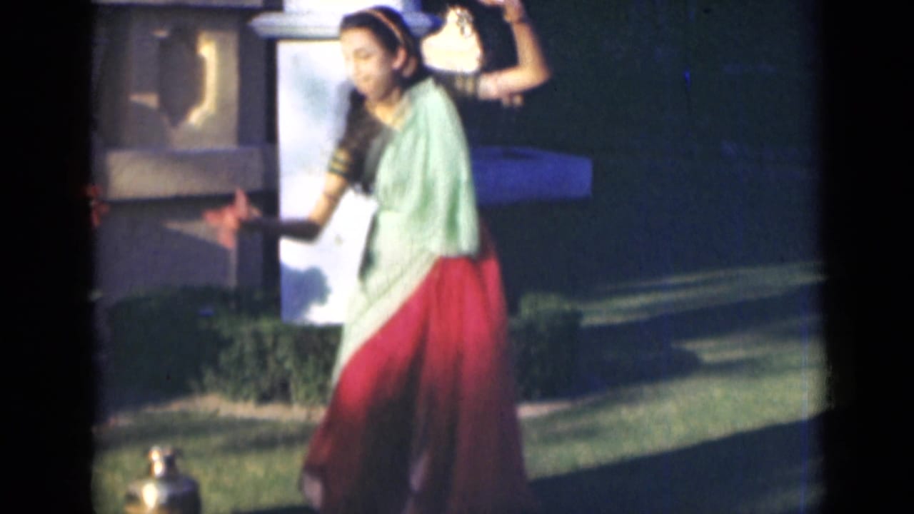 The dancer appears in a pale green blouse and red skirt, extending one arm while performing outside in the India home-movie scene.