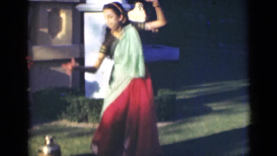 The dancer appears in a pale green blouse and red skirt, extending one arm while performing outside in the India home-movie scene.