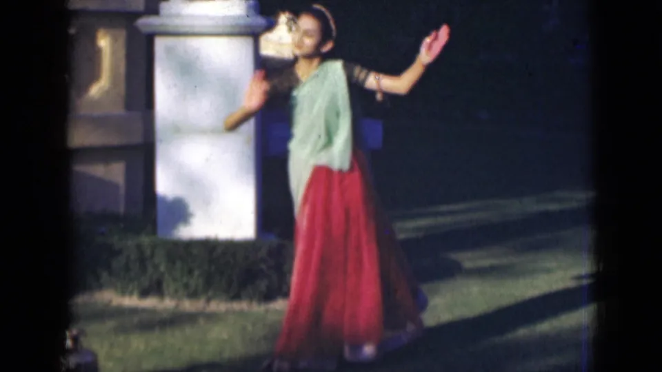 A young woman in 1947 India footage raises both hands in a dance pose, standing on grass near a white garden pillar.