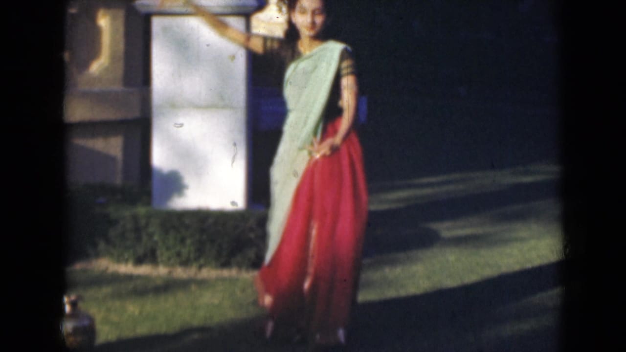 Young woman in a light green and red sari poses beside a white pillar on a lawn.