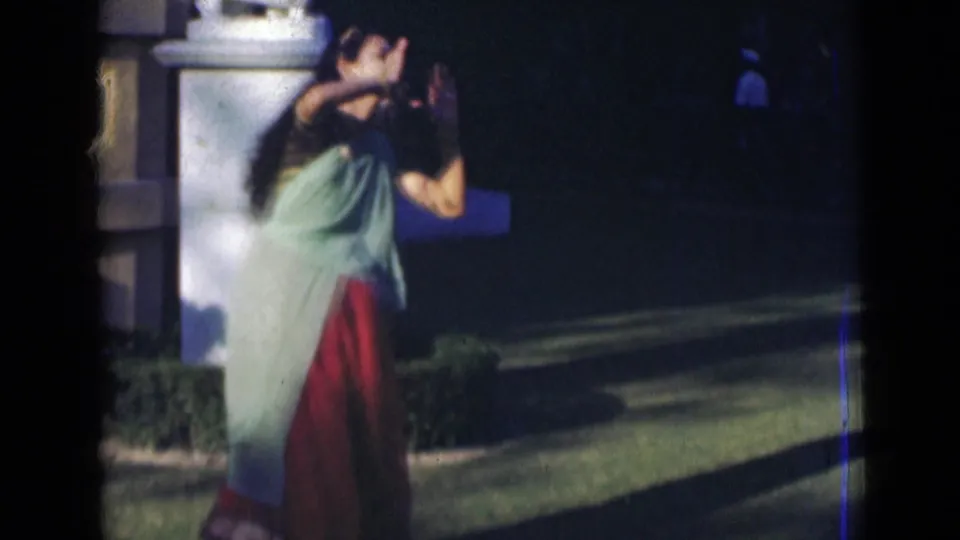 A young woman in a sari bends into an expressive dance movement outdoors, with hedge, lawn, and a distant figure visible in the background.