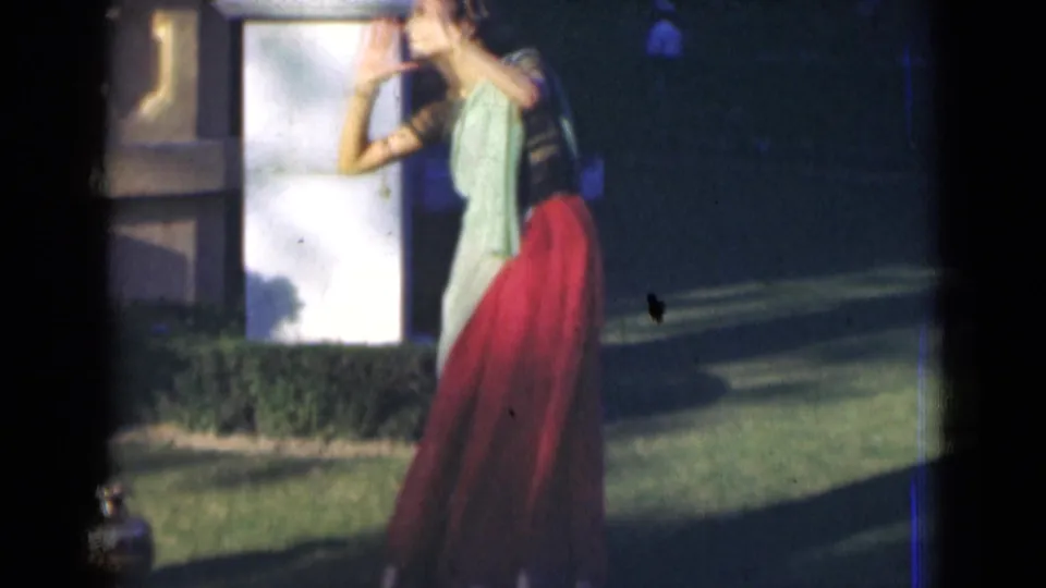 The performer leans sideways in a dance gesture on a lawn in metadata-supported 1947 India, framed by a white pillar and long shadows.